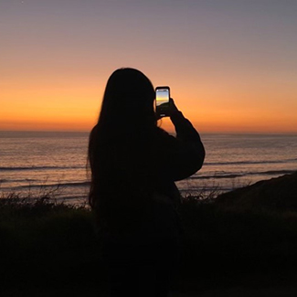 photo taken from behind a person looking out at the ocean at sunset while holding a cell phone taking a picture