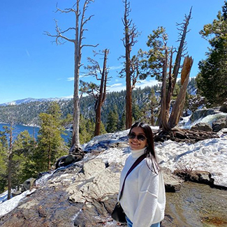 photo of person with long black hair wearing a white turtleneck sweater standing on a mountain with green trees and a body of blue water in the background