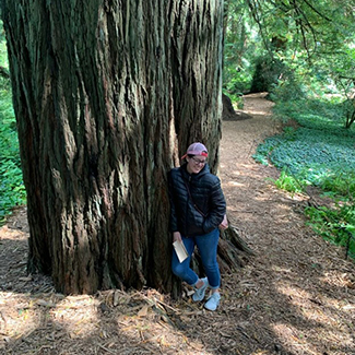 photo of a person wearing blue jeans and a black sweater leaning against a large tree on a forest path with trees and green plants in the background